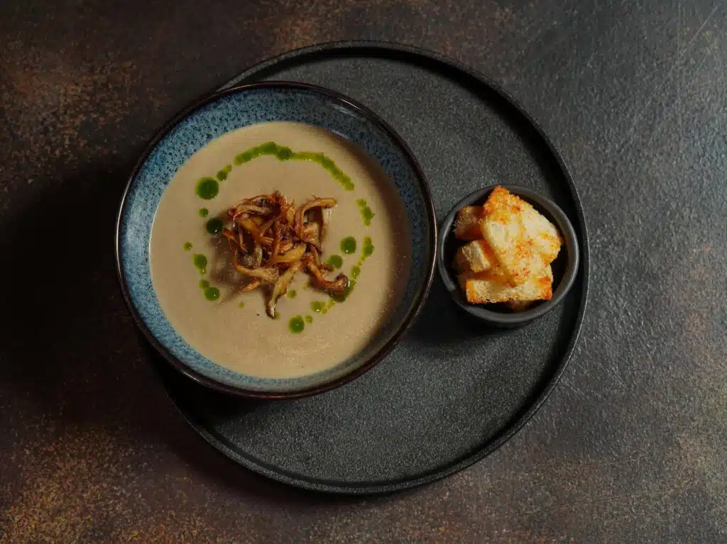 Velouté de champignons, oignons caramélisés et huile de persil dans un bol bleu émaillé, croûtons dorés servis à part, restaurant ARIA Paris